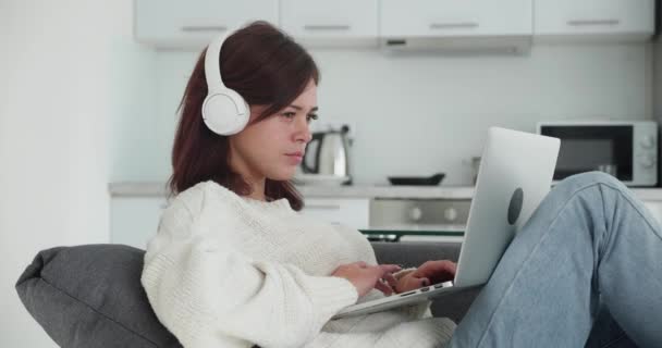 Mujer joven sonriente en auriculares sentados en el sofá usando portátil portátil mirando el mensaje de escritura de pantalla, señora feliz charlando en la computadora navegando por Internet redes sociales estudiando o trabajando — Vídeos de Stock