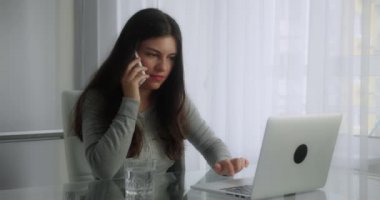 Genç ve meşgul bir esmer kadın masasında oturmuş telefonla konuşuyor ve modern ofis geçmişinin bilgisayarına bakıyor.