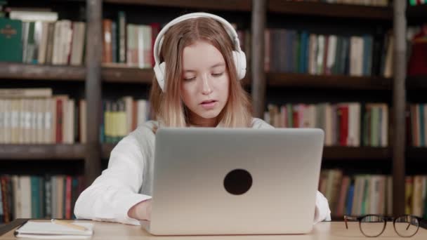 Bibliotheek: Gifted Girl maakt gebruik van laptop, Writes Notes for the Paper, Essay, Study for Class Assignment. Leren, studeren voor het examencollege. Bekijk voorkant Portret met boekenplanken — Stockvideo