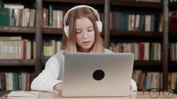 Biblioteca: Garota talentosa usa laptop, escreve notas para o papel, ensaio, estudo para atribuição de classe. Aprendendo, estudando para a faculdade de exames. Vista frontal Retrato com estantes — Vídeo de Stock