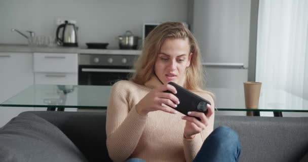 Krullend meisje met blond haar spel met smart phone op moderne keuken achtergrond. Vrije tijd in acht nemen — Stockvideo