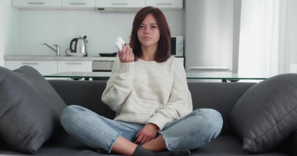 Femme ennuyée regarde un film ennuyeux à la télévision et change de chaînes sur la télécommande se trouve jambes croisées sur le canapé confortable sur fond de cuisine. Fatigué de longues publicités — Video