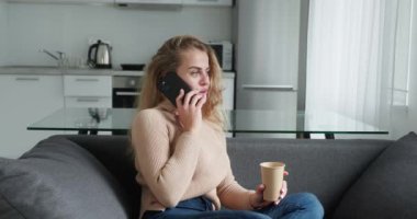 Sarışın kadın cep telefonuyla konuşuyor ve evinin penceresinin yanında oturmuş kahve içiyor.
