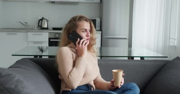 Ξανθιά γυναίκα μιλώντας στο κινητό τηλέφωνο και πίνοντας καφέ, ενώ κάθεται κοντά στο παράθυρο στο σπίτι — Αρχείο Βίντεο