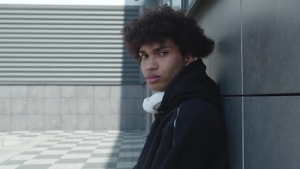 A troubled teenager with a nose piercing in a black hoodie and headphones sits leaning against a concrete wall. Millennial looks thoughtfully at the camera and turns his head to the side — Stock Video