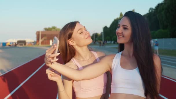 Twee mooie zorgeloze meisjes die selfie nemen met een smartphone. Vrouwen, schattige gezichten. Positieve modellen poseren op stadion achtergrond — Stockvideo