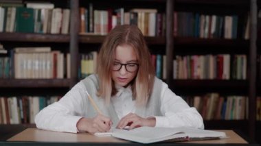 Liseli kız kütüphanedeki bir kitaptan notlar alıyor. Masada oturan beyaz bir kadın kütüphanede ödevlerini yapıyor..