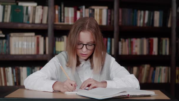 Scolaretta prendere appunti da un libro in biblioteca. Giovane donna caucasica seduta a tavola a fare incarichi in biblioteca. — Video Stock