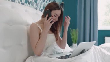 Yatakta pijamalı güzel bir kız telefonda konuşuyor ve dizüstü bilgisayarla çalışıyor.