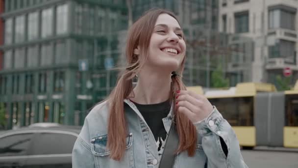 Ein nettes Mädchen in Jeansjacke mit Pferdeschwanz richtet vor der Kulisse der Stadt ihre Haare vom Wind. Jahrtausendelanges Weibchen unbeschwert — Stockvideo