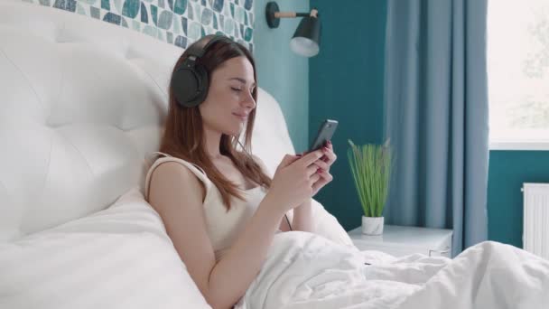 Ontspannen jonge brunette vrouw in hoofdtelefoon liggend op wit bed met smartphone in handen. Aantrekkelijk meisje ontspannen thuis met moderne gadget. — Stockvideo