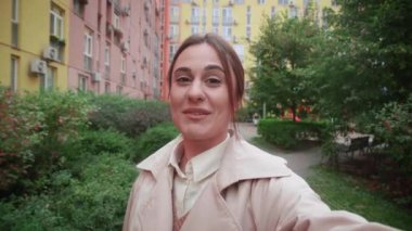 Gülümseyen hoş bir kadın merhaba jesti, elinde kamera, telefon görüşmesi, iş arkadaşları veya Freands ile iletişim. Güzel kadın blogcu şehrin arka planında video kaydediyor