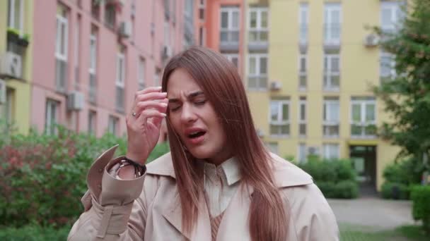 스트레스를 받은 젊은 여성의 얼굴에 두 통 이있는 베이지 코트를 입은 모습. 화려 한 건물 근처 야외에서 있는 모습 — 비디오
