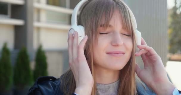 Klidná studentka ve sluchátkách poslouchat hudbu a dívat se do kamery stojící venku. Létající blond vlasy ve větru. Millennial blondýny dívka mají volný čas. — Stock video