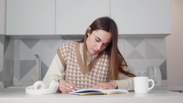 Young woman with expression reads a book while sitting at a table in the kitchen — Stock Video