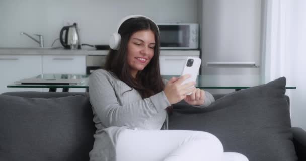 Morena jovem mulher tem vídeo chat no smartphone sentado em casa sobre o fundo da cozinha — Vídeo de Stock