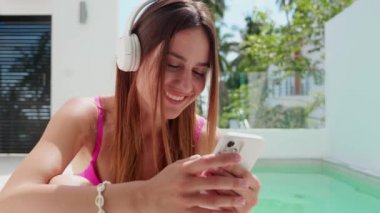 Kulaklıklı, mayo giymiş bir kızın yakın plan fotoğrafı. Telefonu havuzun arka planında tut..