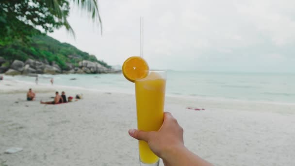 Kvinna hand hålla nypressad apelsinjuice med havet linje och stenar på en bakgrund. — Stockvideo