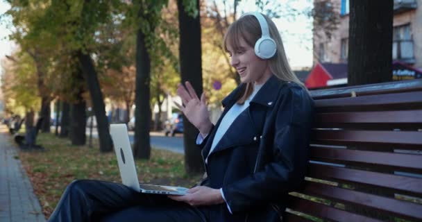 Glad ung kvinna med hörlurar som använder en bärbar dator i parken. Blond kvinna student har videosamtal med vänner eller föräldrar — Stockvideo