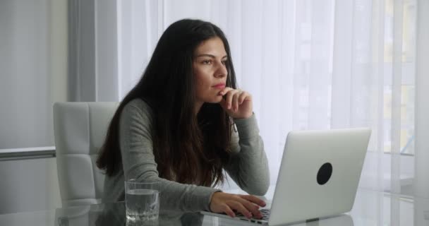 A young brunette looking for ideas for a scientific article. Female sitting at a table looking to the side and at the monitor screen, typing a dissertation on a laptop — Stock Video
