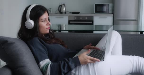 ブルネットの女性は自宅のオフィスでソファに横たわっているラップトップのウェブカメラ会議の仕事を使用してリモートジョブインタビューのためのビデオ通話を持っています。幸せな女性は、仮想チャット会議で話しています日付オンラインコンピュータを使用して — ストック動画