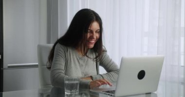 Uzun saçlı esmer kadın, mutfak arka planında otururken bilgisayarında görüntülü konuşma yapıyor. Kadın, ekrana bakarken elini sallayarak gülümsüyor.