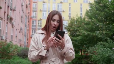 Açık havada duran bej ceketli bir kadın. Gözlerime inanamıyor. Akıllı telefon kullanıyor, gülümsüyor ve şaşırmış görünüyor..