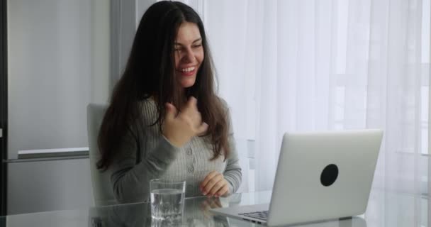 Esmer, bin yıllık uzun saçlı bir kadın mutfak arka planında otururken bilgisayarında görüntülü konuşma yapıyor. Kadın, ekrana bakarken elini sallayarak gülümsüyor. — Stok video