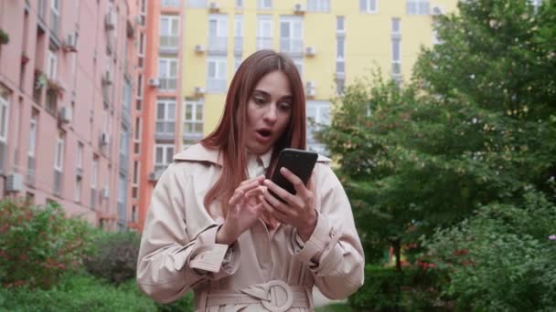 Femme en manteau beige debout à l'extérieur. Elle ne peut pas croire mes yeux en utilisant un smartphone, souriant et regardant surpris, disant wow. — Video