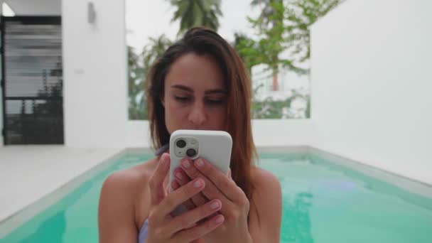 スマートフォン付きのヴィラでプールで美しい若いビキニの女性。プールのビキニの女の子は、携帯電話でテキストメッセージを使用して。旅行者の女性の笑顔観光幸せな休日の美しさ休暇夏 — ストック動画