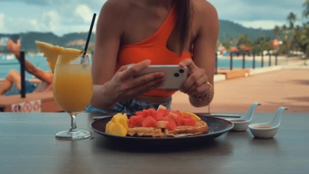 Female blogger photographing orange juice in cafe with her phone. A young woman taking photo waffles on smartphone, photographing meal with mobile camera. — Stock Video