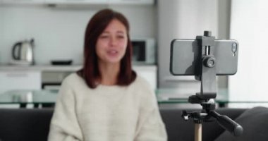 Süveter giymiş genç bir video blogcusunun akıllı telefondaki vlog 'u kaydederken kameraya konuştuğu çekimler tripoda sabitlendi