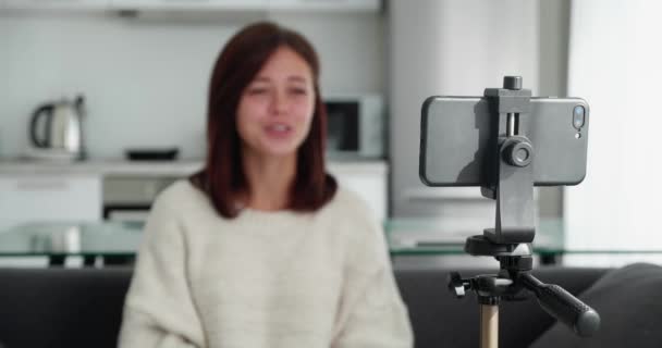 Tracking shot with rack focus of young female video blogger in a sweter talking to camera when recording vlog on smartphone fixed on tripod — Wideo stockowe