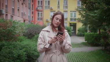 Açık havada duran bej ceketli bir kadın. Akıllı telefon kullandığım için gözlerime inanamıyor, kötü haberlerden rahatsız oluyor..