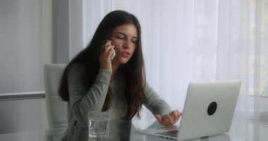 Genç ve meşgul bir esmer kadın masasında oturmuş telefonla konuşuyor ve modern ofis geçmişinin bilgisayarına bakıyor.
