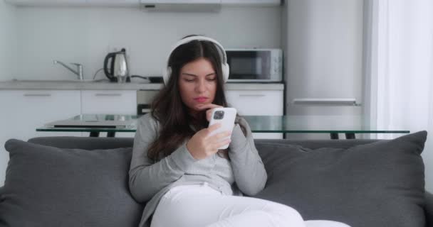 Šokovaná brunetka dívka v šedém svetru říká ano hledá smartphone a zakrývá ústa, zatímco sedí v gauči kuchyně pozadí — Stock video