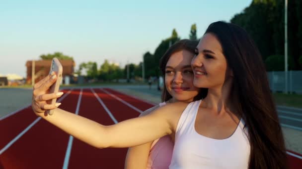 Dos hermosas chicas jóvenes en ropa deportiva se están tomando una foto selfie usando un teléfono inteligente en el fondo del estadio — Vídeos de Stock