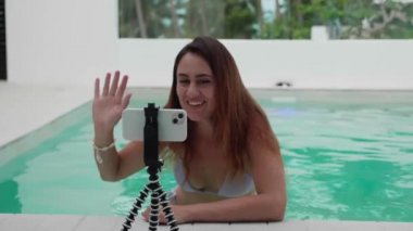 Gülümseyen genç bir kadın, akıllı telefondan havuzda tripoda sesleniyor. Seyyar vlog çeken kadın blogcu, canlı yayın, çevrimiçi konsept arıyor