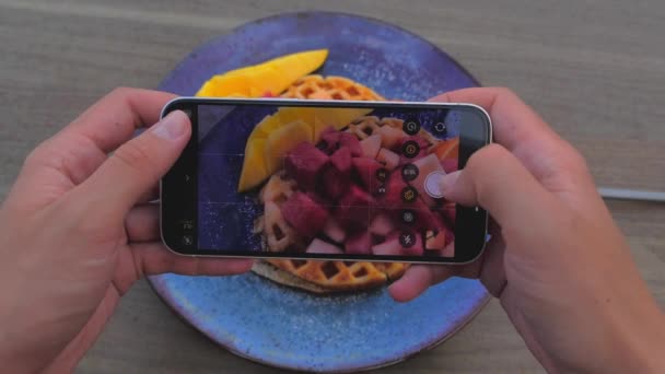 Жіночі руки використовують смартфон, щоб сфотографувати смачну вафельку з червоними фруктами та манго — стокове відео