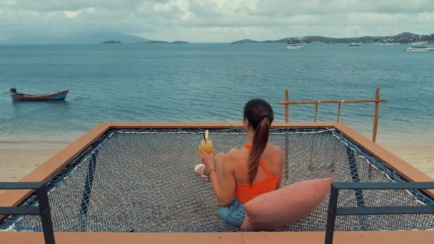 Bela jovem mulher sentada na rede beber coquetel ou suco de laranja na praia do mar. menina sexy, relaxamento, conceito de férias — Vídeo de Stock