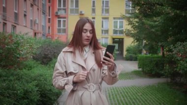 Şok olmuş esmer bir kadın, bej ceketli, akıllı telefonuyla 