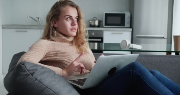 Wanita pirang yang cantik memakai headphone duduk di tempat kerjanya di rumah ketika berpikir tentang jawaban ketika berbicara dengan teman-teman di media sosial — Stok Video