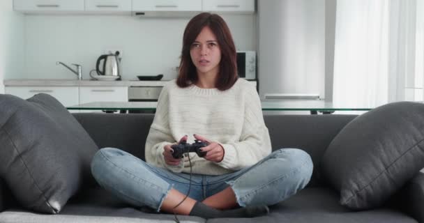 Bela jovem gamer animado sentado em um sofá, jogando e ganhando em jogos de vídeo em um console. Ela joga com um controle sem fio. Quarto acolhedor é iluminado com luz quente e néon. — Vídeo de Stock