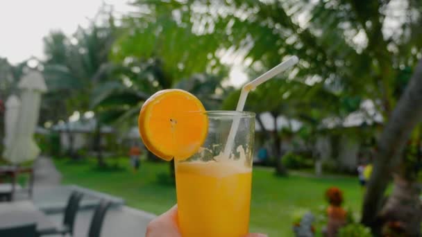 Närbild kvinnlig hand hålla nypressad apelsinjuice med mot bakgrund av exotiska gröna träd på bakgrunden. — Stockvideo