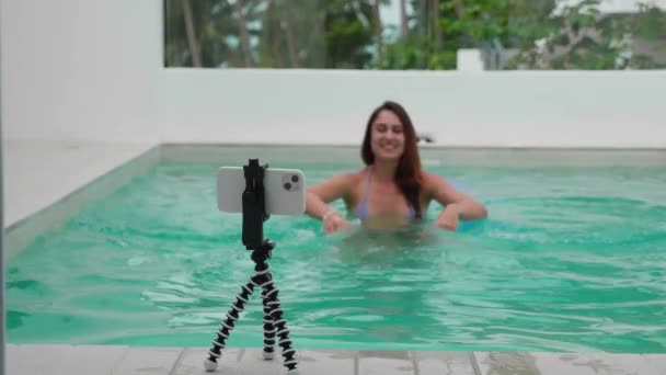 Молода дівчина-блогер записує на камеру для контенту соціальних мереж, використовуючи телефон на тринозі на відкритому повітрі, крупним планом. Вона спілкується в басейні . — стокове відео