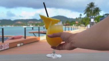 Bir bardakta alkolsüz ananas kokteyli ile tropikal bir plaj arka planında el ele tutuşmak.