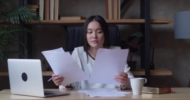 Серьёзная молодая азиатская бизнесвумен проверяет корпоративную переписку, сидя за столом домашнего офиса. Женщина-предприниматель читает документы, анализирует финансовые документы, готовит аудиторский отчет на — стоковое видео