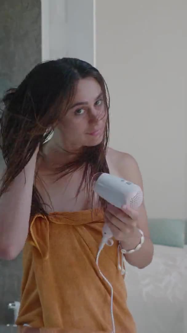 Jonge mooie vrouw staat voor de spiegel en met behulp van haardroger tijdens het drogen van haar na het baden — Stockvideo