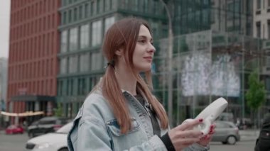 Kot ceketli genç, günlük giysiler içinde şehir merkezinde yürüyor ve kablosuz kulaklık kullanarak ses kitabı dinliyor.