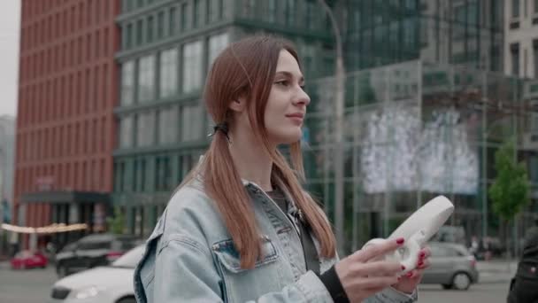 Adolescente en una chaqueta de mezclilla con ropa de abrigo casual caminando en el centro y escuchando audiolibros usando auriculares inalámbricos — Vídeo de stock